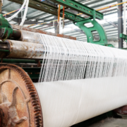 Industrial weaving machine in a textile factory, producing white fabric with visible machinery parts and threads.