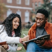 Two students sitting on grass, studying together, one writing in a notebook and the other holding an open book, smiling.