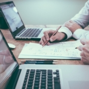 Two people working at a desk with laptops, reviewing documents and taking notes with pens.